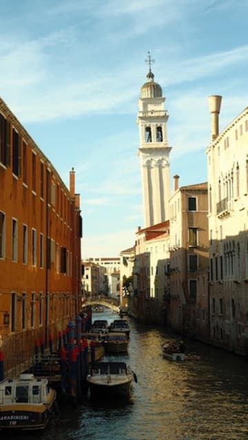 A tall white bell tower rising above a narrow canal lined with boats and old buildings