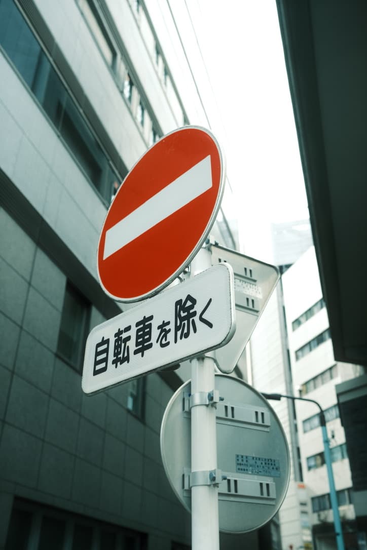 A Japanese no-entry road sign with a plate reading 自転車を除く, framed by office buildings
