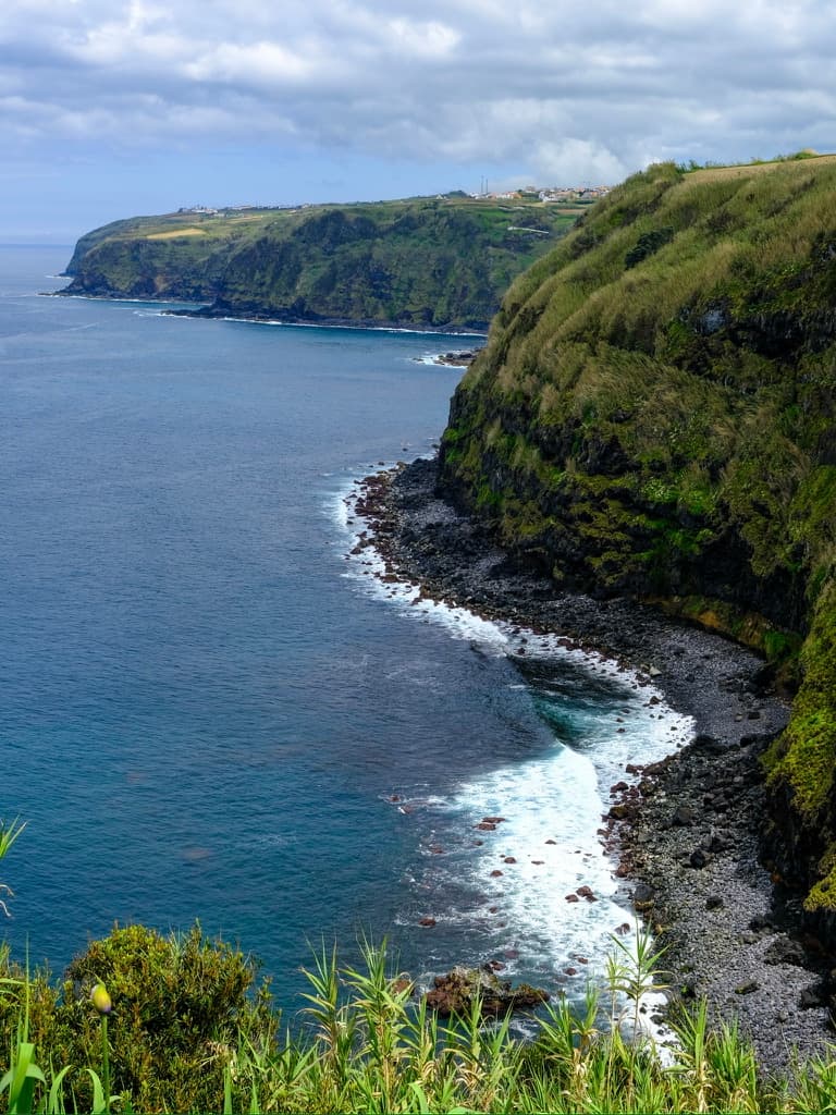 Surf breaking on black volcanic rocks beneath tall green sea cliffs
